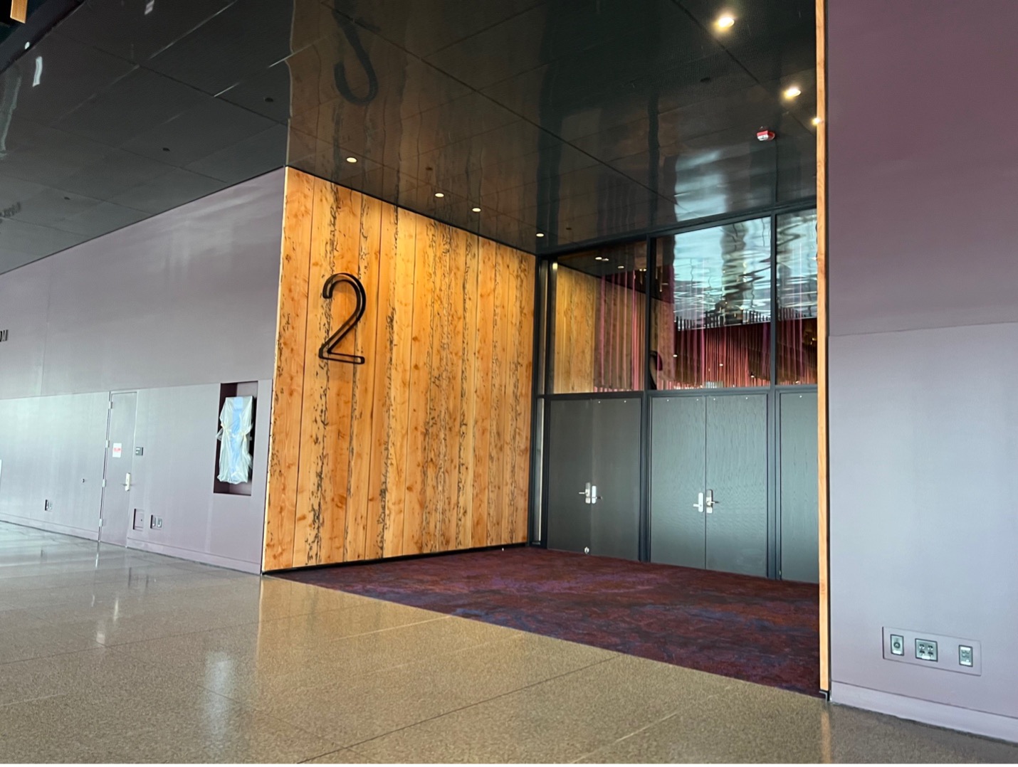 The entrance to Ballroom 2 is located on the right side of the main hallway. Each ballroom has an alcove with purple carpeting and walls with wooden panels. Each ballroom has a large cutout on the wall with the number of the ballroom. The doors in this photo to the ballroom are black.