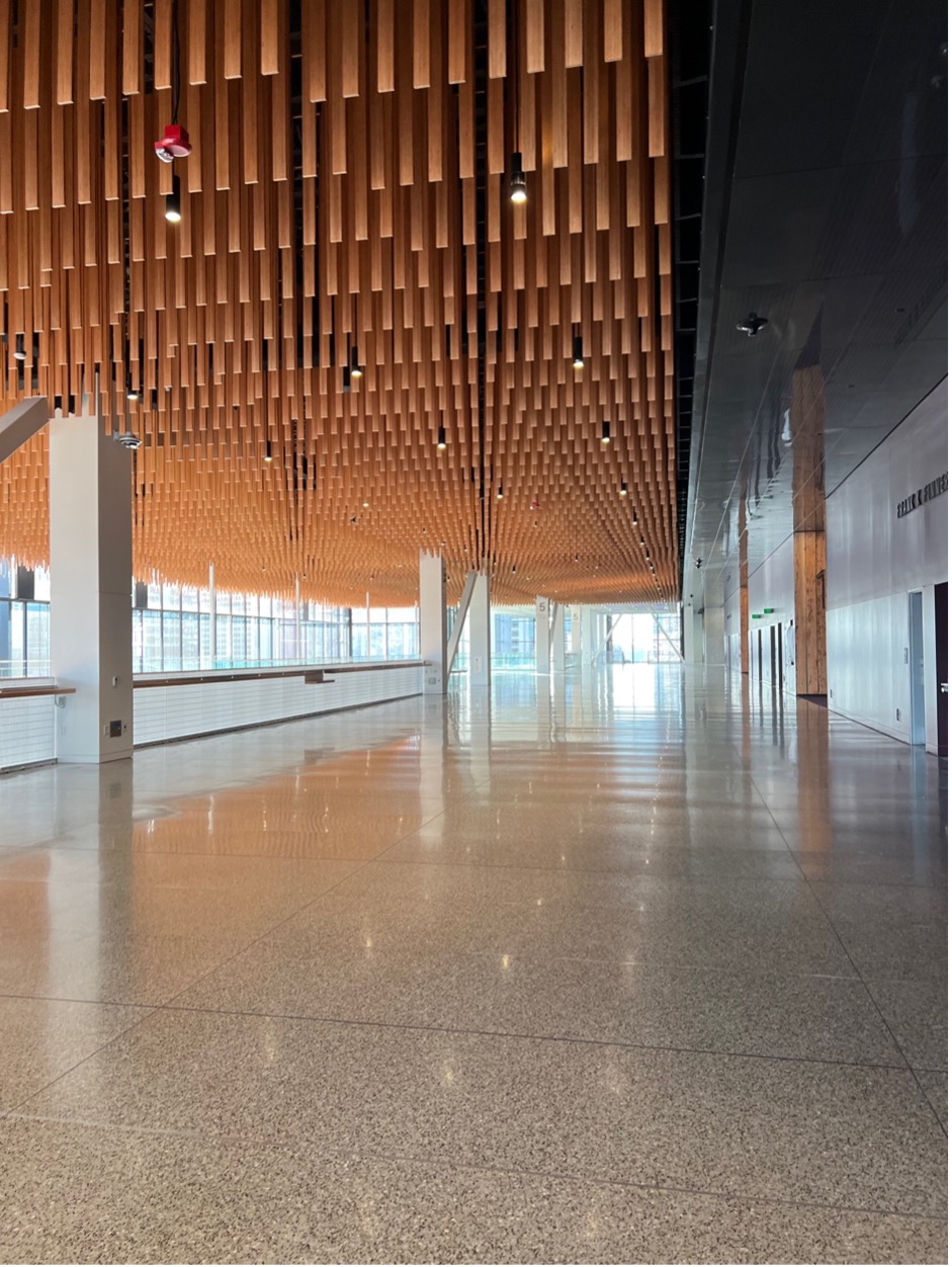 A long hallway with speckled gray flooring. To the left is open to below with large glass windows. From the ceiling are hung wooden slats.