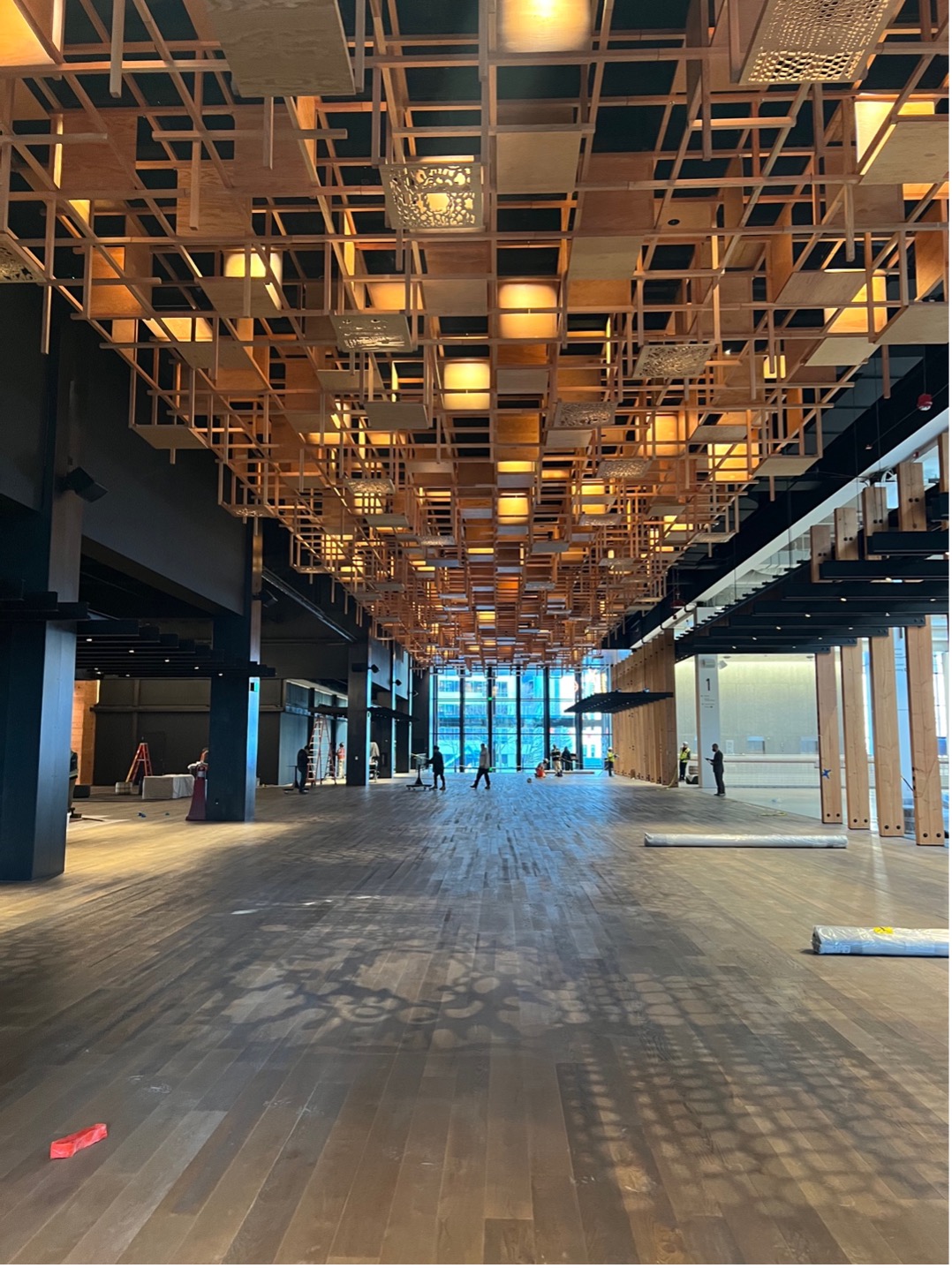A photo of the main entry hallway. The ceiling is a wood panel design. The floor is a light brown wood paneling. Down the hallway about halfway, the hallway opens to the right and to the left. The wooden ceiling design casts branch-like shadows on the wood flooring.