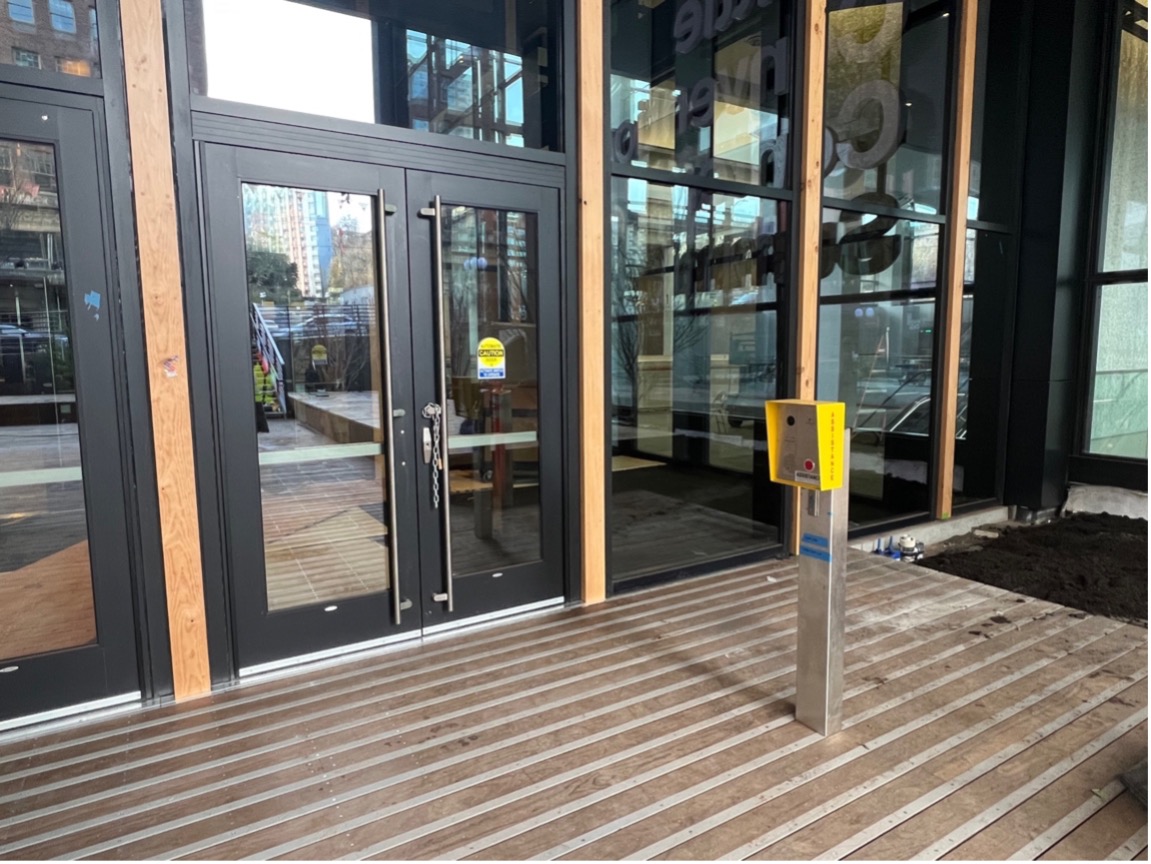 This photo shows the right most double doorway. It is marked as for accessibility. To the right of the door is a yellow call box on a silver post. On the silver post is a wheelchair button. On the callbox is a red button to press if needing access during off-hours.